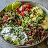 A vibrant, close-up view of a healthy taco bowl featuring seasoned ground beef, crisp romaine lettuce, and a zesty lime yogurt crema drizzle.