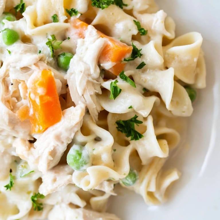 Savory shredded chicken and wide egg noodles with carrots and peas in a light creamy sauce, garnished with parsley in a warm skillet.